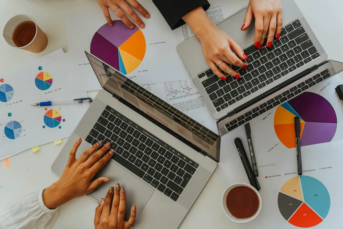 Overhead view of two analysts typing on laptops surrounded by colorful pie-chart printouts
